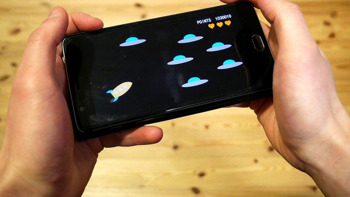 Two hands holding a smartphone sideways, playing a simple game with a rocket and flying saucers on screen
