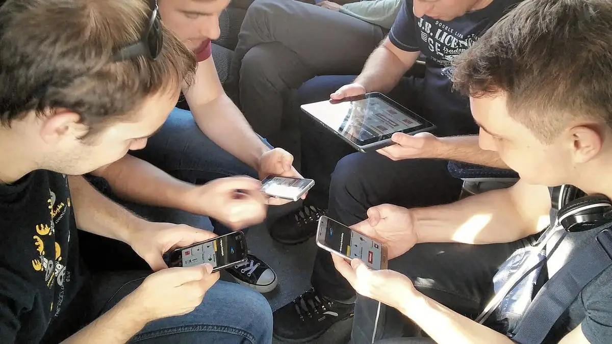 Four adults sit in a close circle outdoors, each holding a phone or a tablet running the same multiplayer game