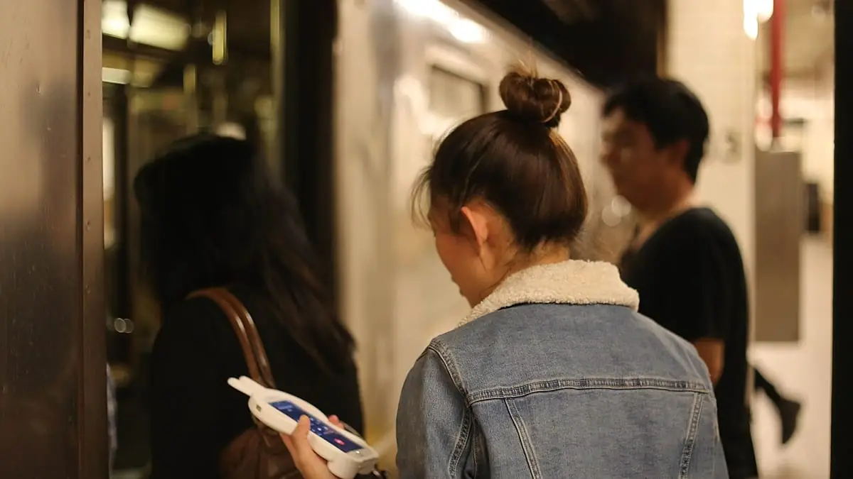 A transit rider in a denim jacket holds a smartphone with its screen lit, standing beside a subway train among other passengers